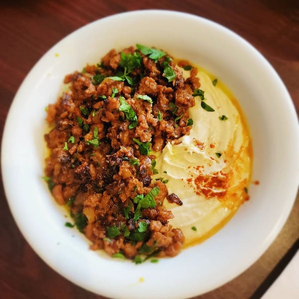 A white bowl of creamy hummus topped with seasoned ground meat, fresh parsley, and paprika.