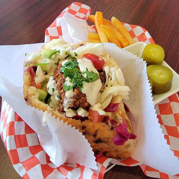 A falafel pita sandwich with fries and pickles, served in a red and white checkered paper.