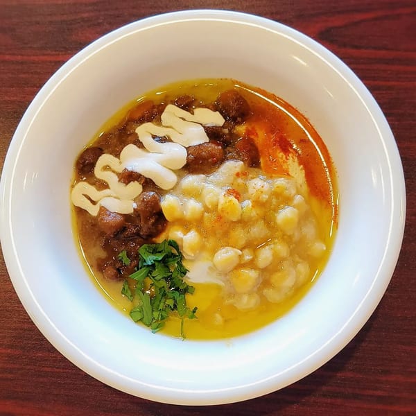 A white bowl of Hummus Yummy's Mediterranean dish with chickpeas, meat, sauce, and parsley.